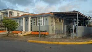 Um edifício de dois andares com telhado plano, varanda coberta e um jardim frontal cercado.