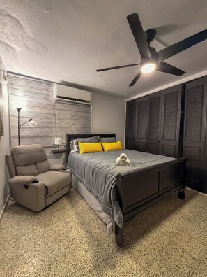 A bedroom with a bed, a reclining chair, a ceiling fan, and an air conditioning unit.