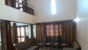 A spacious living room with a high ceiling, a ceiling fan, a large window, and a view of the outside. There are several wooden chairs and a sofa set. - Aashvi Homes - 4BHK family friendly apartment (JAIPUR)