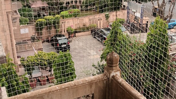 A view through a wire mesh balcony with a garden in the foreground, a parking area, and buildings in the background. - Aashvi Homes - 4BHK family friendly apartment (JAIPUR)