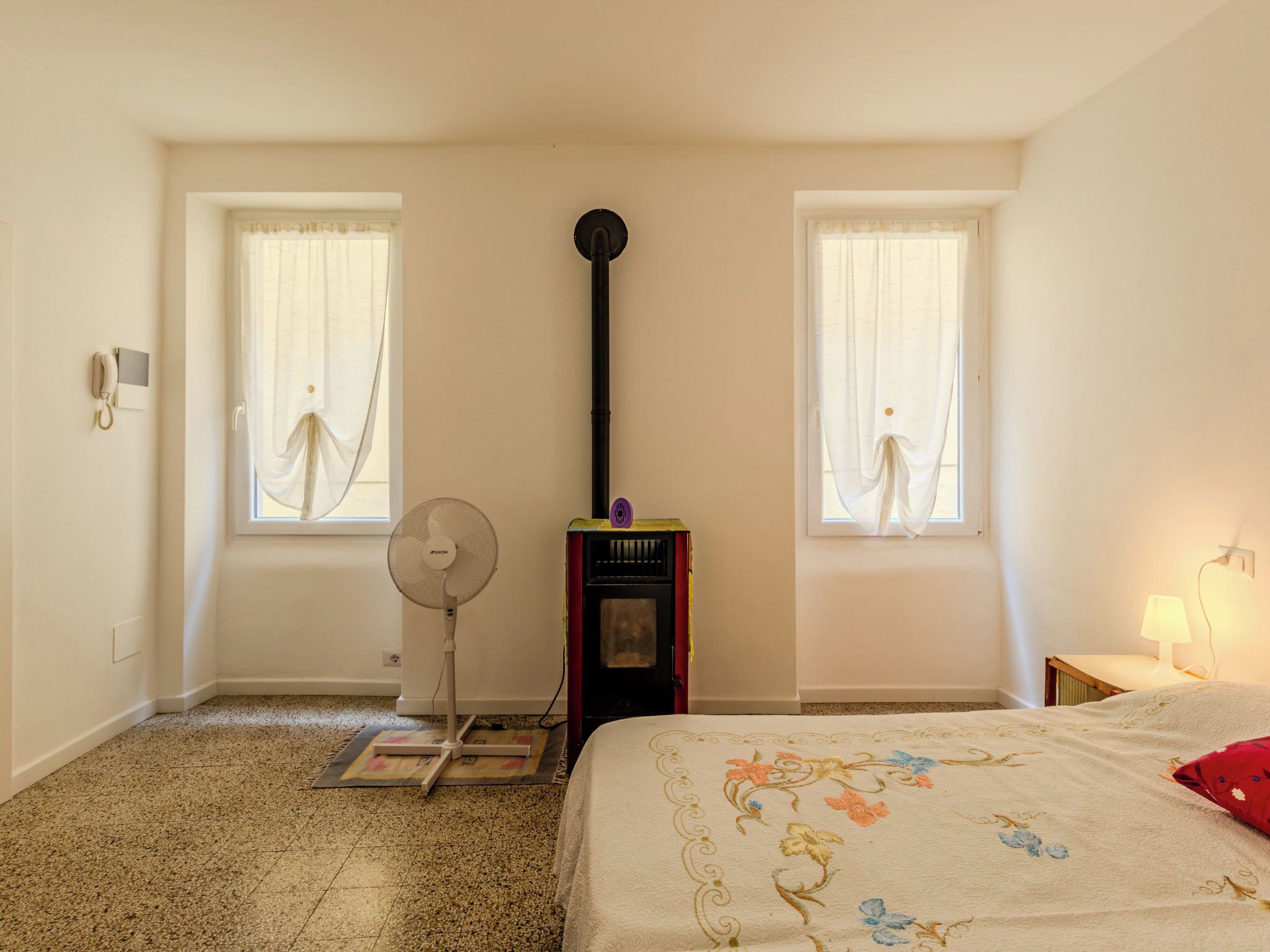 Ein Schlafzimmer mit zwei Fenstern, einem Standventilator, einem Heizgerät und einem Bett mit floralem Überwurf.