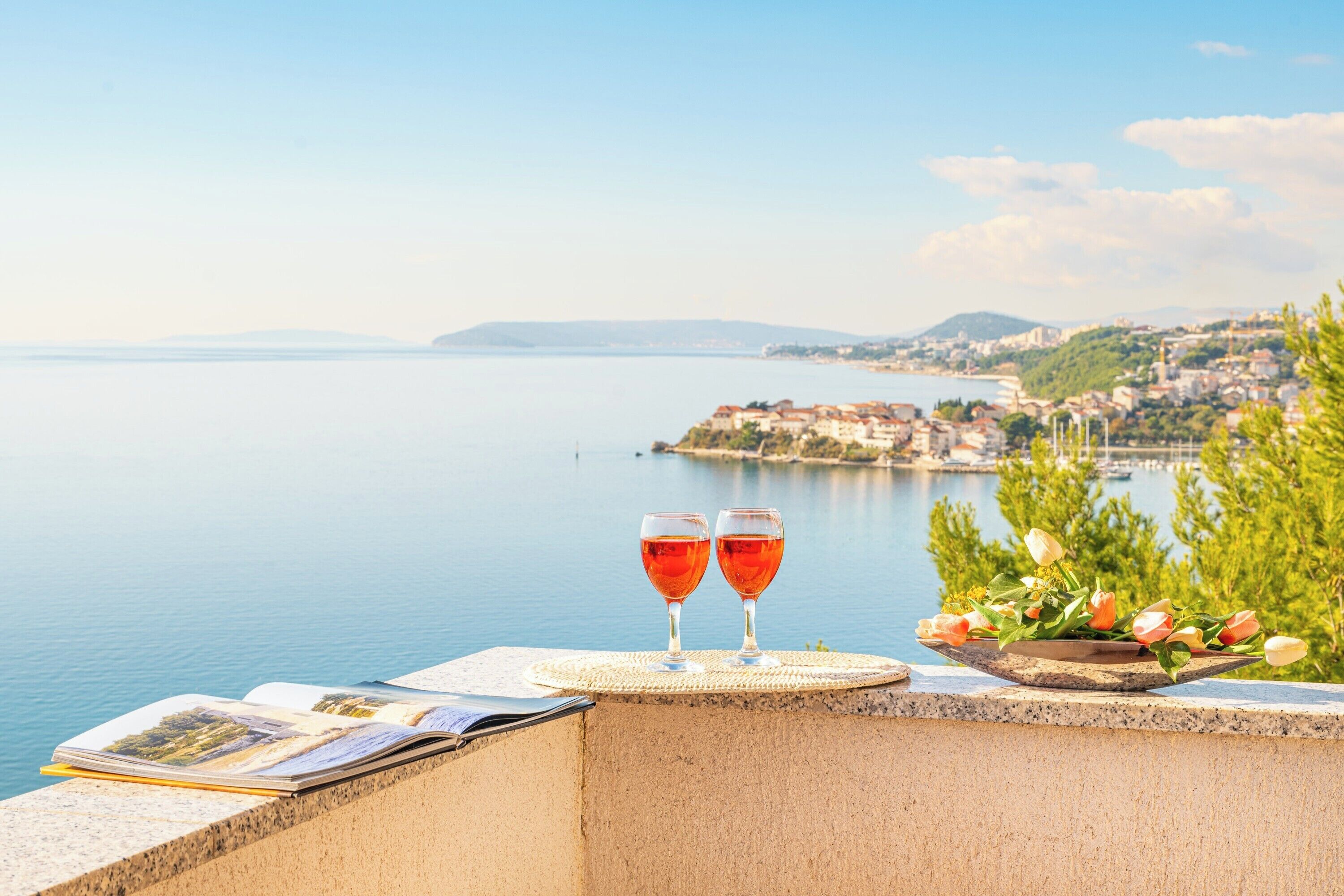 A seaside view with two glasses of red wine on a balcony overlooking calm waters and a distant town.