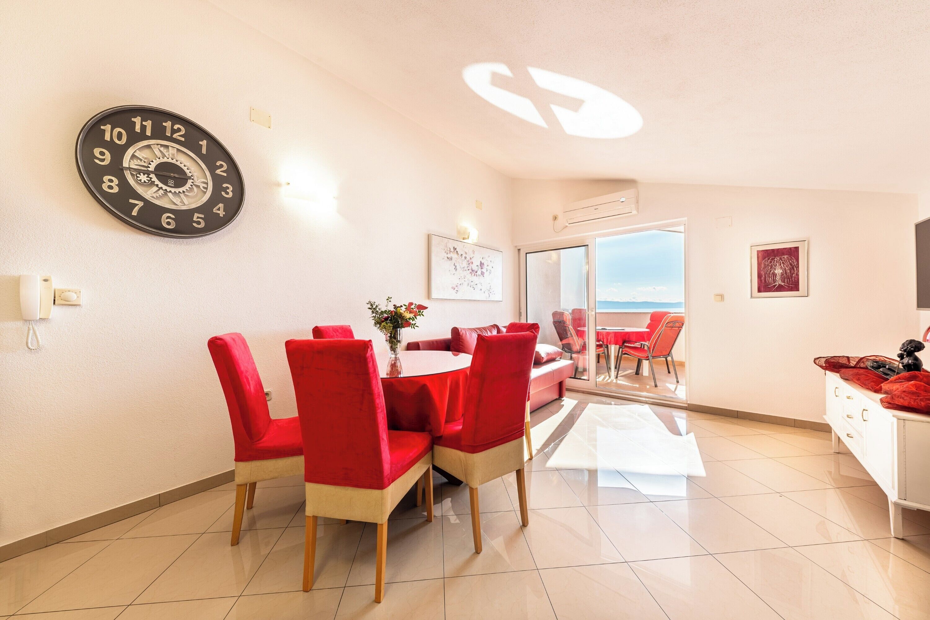 Un coin repas avec une table ronde, des chaises rouges, une horloge murale et une vue sur la mer, visible à travers une porte vitrée.