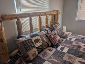 Un lit avec une couette ornée de motifs animaliers, une tête de lit en bois et une fenêtre avec des stores.