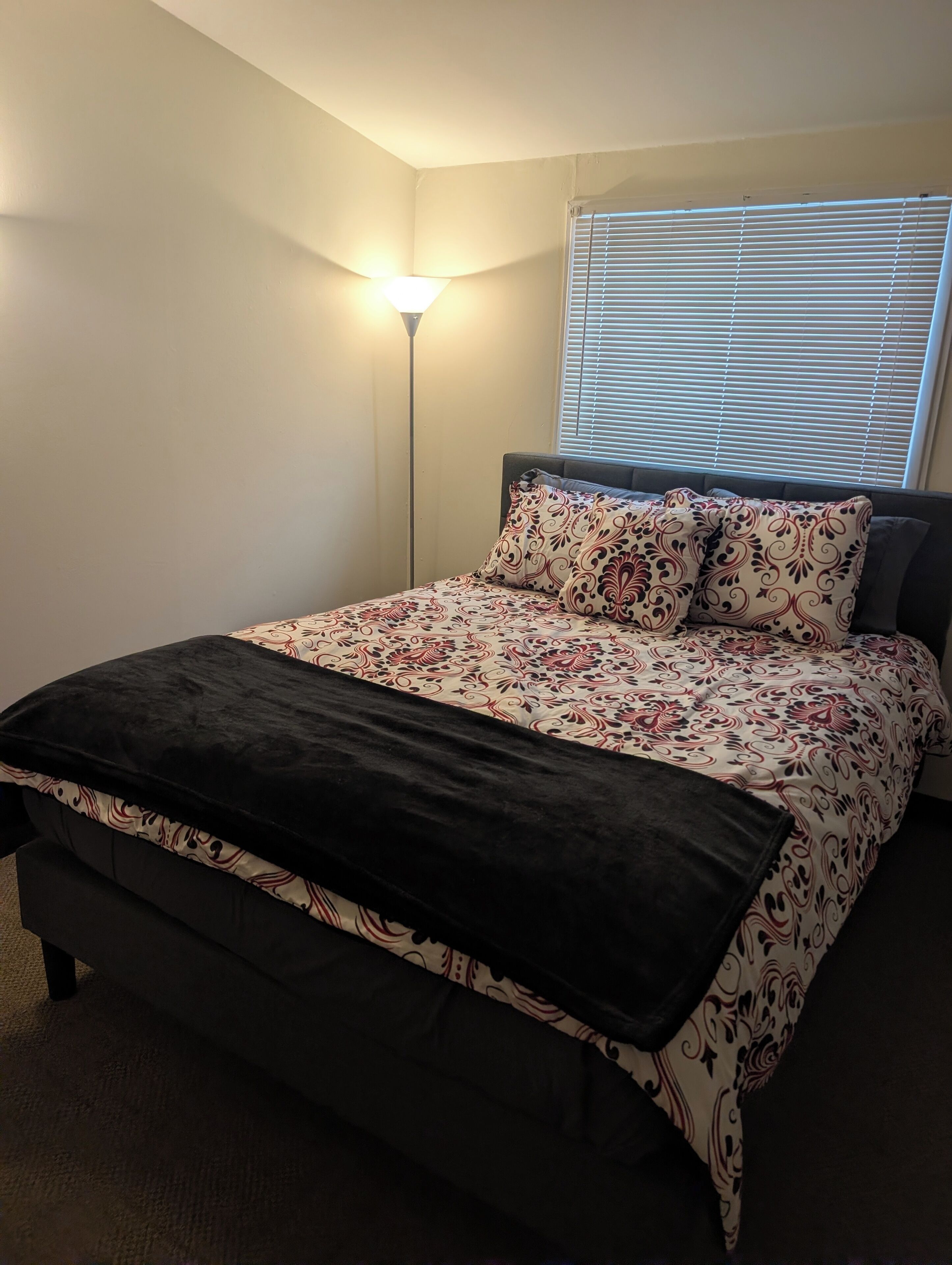 A bedroom with a bed, a floor lamp, and a window with blinds.