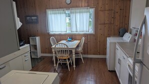A compact kitchen with wooden panel walls, a dining table with chairs, a clock, and a window with curtains. - Near the park! 3-bedroom house with wifi and AC (White Pine)