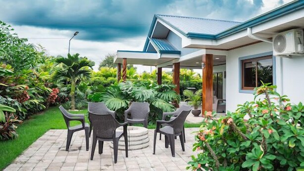 A covered patio with outdoor furniture, surrounded by lush greenery and a house with a blue roof.