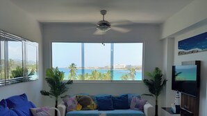 Un salon comprenant un canapé, une table basse, un téléviseur et une grande fenêtre donnant sur la mer.