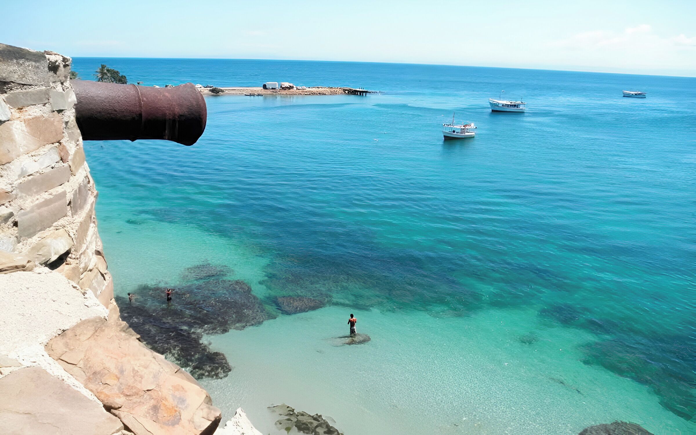 Un paysage côtier avec une eau turquoise et limpide, un mur de pierre et un vieux canon rouillé.