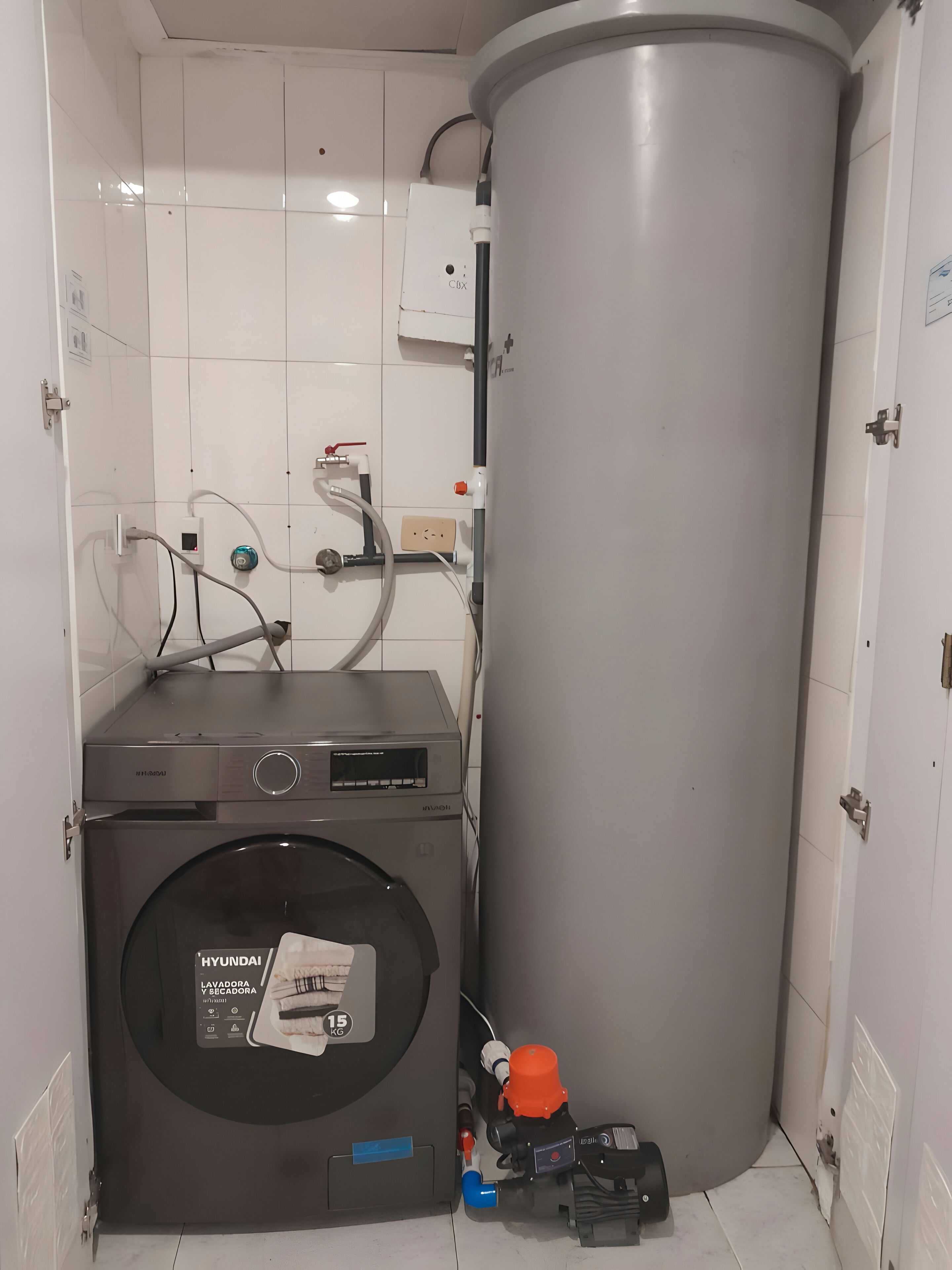 Une petite buanderie équipée d’un lave-linge à chargement frontal, d’un grand chauffe-eau cylindrique et de tuyauteries apparentes.