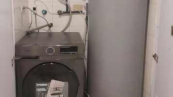Une petite buanderie équipée d’un lave-linge à chargement frontal, d’un grand chauffe-eau cylindrique et de tuyauteries apparentes.