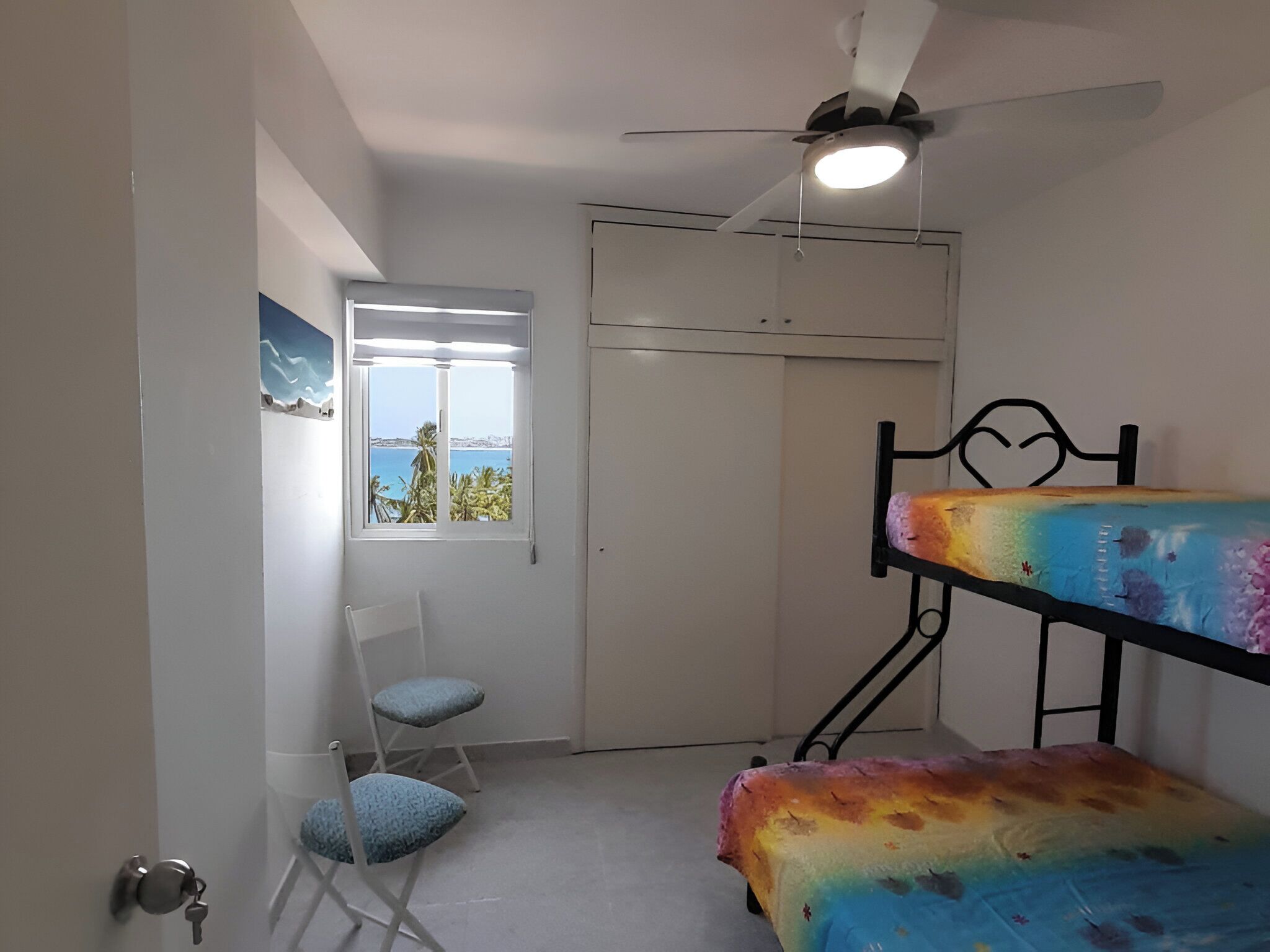 Une chambre avec un lit superposé, une chaise, une porte avec une clé, un ventilateur de plafond et une fenêtre donnant sur la mer.
