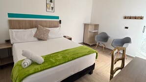 A modern hotel room with a large bed, a green throw blanket, a neatly folded towel, a wooden headboard, a wall-mounted artwork, a wooden desk, and two chairs. - Hotel casa Vecchia (Cali)