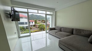 A modern living room with a large grey sofa, a flat-screen TV mounted on the wall, and a sliding glass door leading to a balcony with a view of buildings and greenery. - Amazing 4-bedroom third floor apartment in San Ramón (San Ramón)