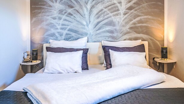 A neatly made bed with white and dark pillows, a quilted blanket, and a wall with a textured design. There are two bedside tables with lamps and a power outlet. - Adelsberg Inn by Interhome (Postojna)