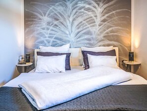 Un lit bien fait, avec des oreillers blancs et foncés, une couverture matelassée et un mur au motif texturé. On y trouve deux tables de chevet avec des lampes et une prise de courant.