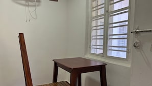 A corner room with a wooden chair and table, a window with bars, and a wall-mounted power outlet.
