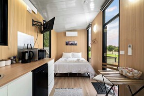 A compact room with a bed, a small kitchenette, a television, and a window offering a view of the outdoors.