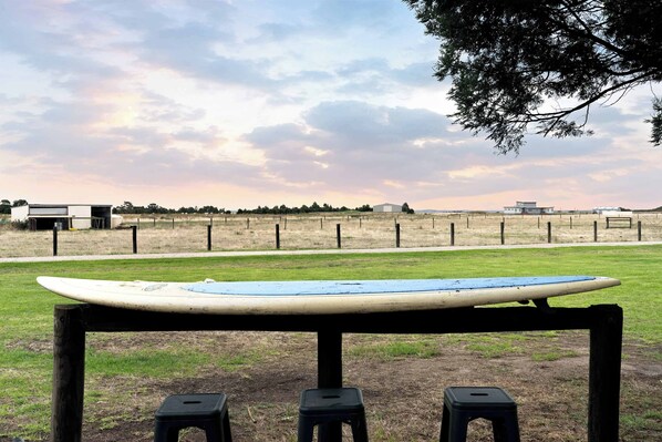 Una tabla de surf sobre un soporte, dos taburetes, un campo cubierto de césped, una cerca y un edificio al fondo.