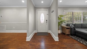 A hallway with a white door, wooden flooring, a grey couch, and a window with blinds.