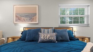 A bedroom with a large bed, a framed painting of a beach, a window with blinds, and a bedside table with a lamp.