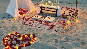 Montagem de praia com uma tenda, uma mesa de jantar com velas e pétalas de rosa na areia.