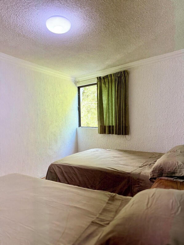 A room with two beds, a window with green curtains, and a textured white wall.