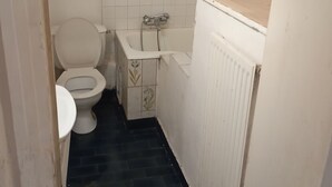 A small, narrow bathroom with a toilet, sink, and shower. The floor is dark, and the walls are tiled.