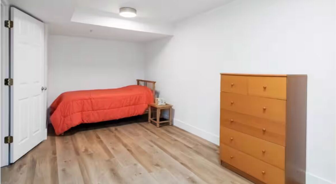 Een slaapkamer met een houten vloer, een bed met een oranje dekbedovertrek, een houten dressoir en een witte deur.