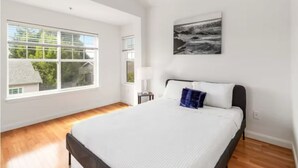 Een slaapkamer met een groot raam, een bed met wit beddengoed, een blauw kussen en een kunstwerk aan de muur.