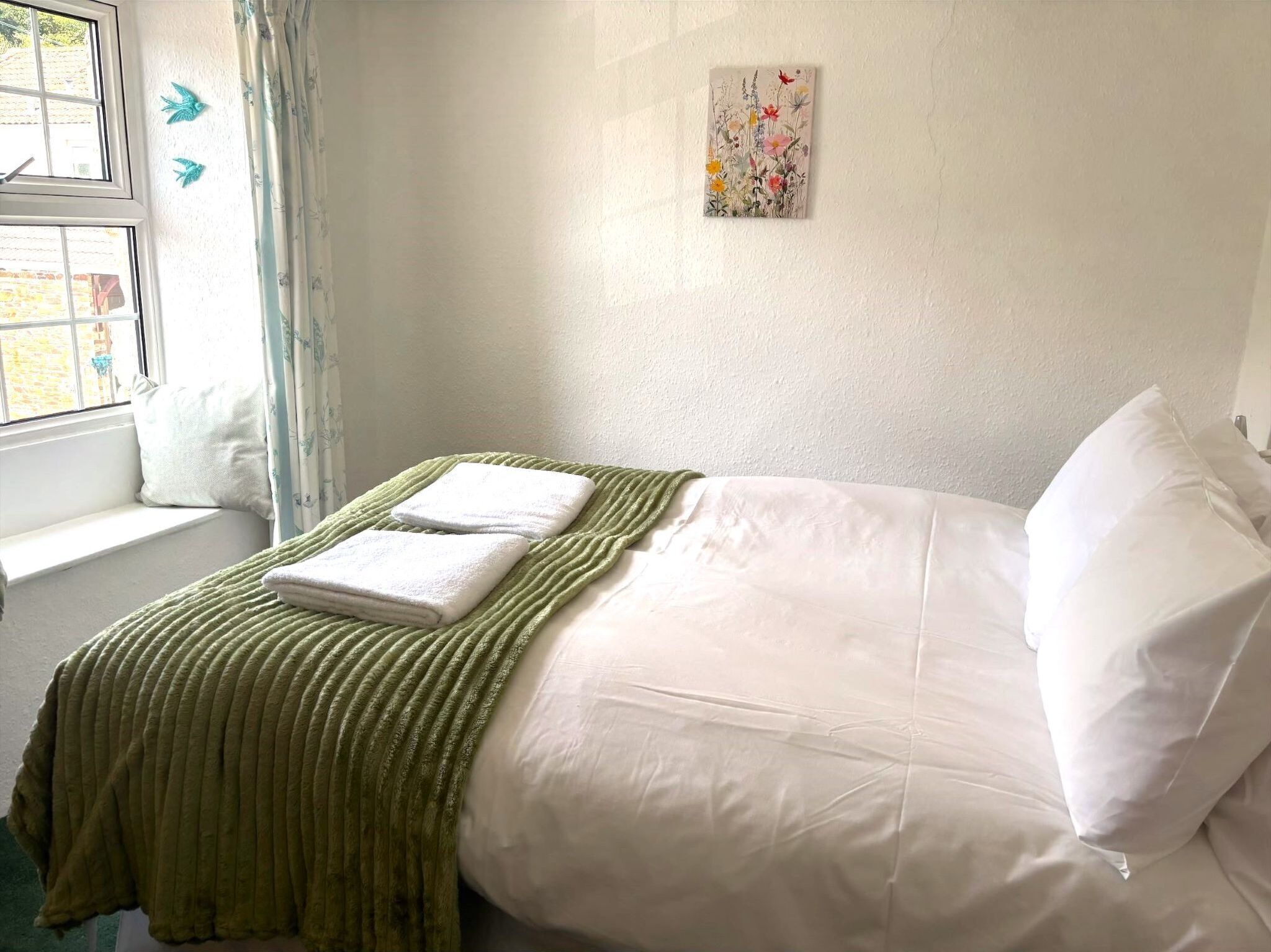 A neatly made bed with white linens and a green throw blanket, a window with curtains, and a wall-mounted artwork.