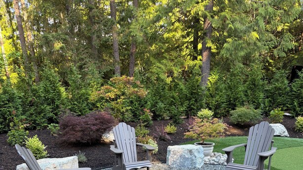 An outdoor seating area with Adirondack chairs and a fire pit, surrounded by a gravel ground and lush greenery.