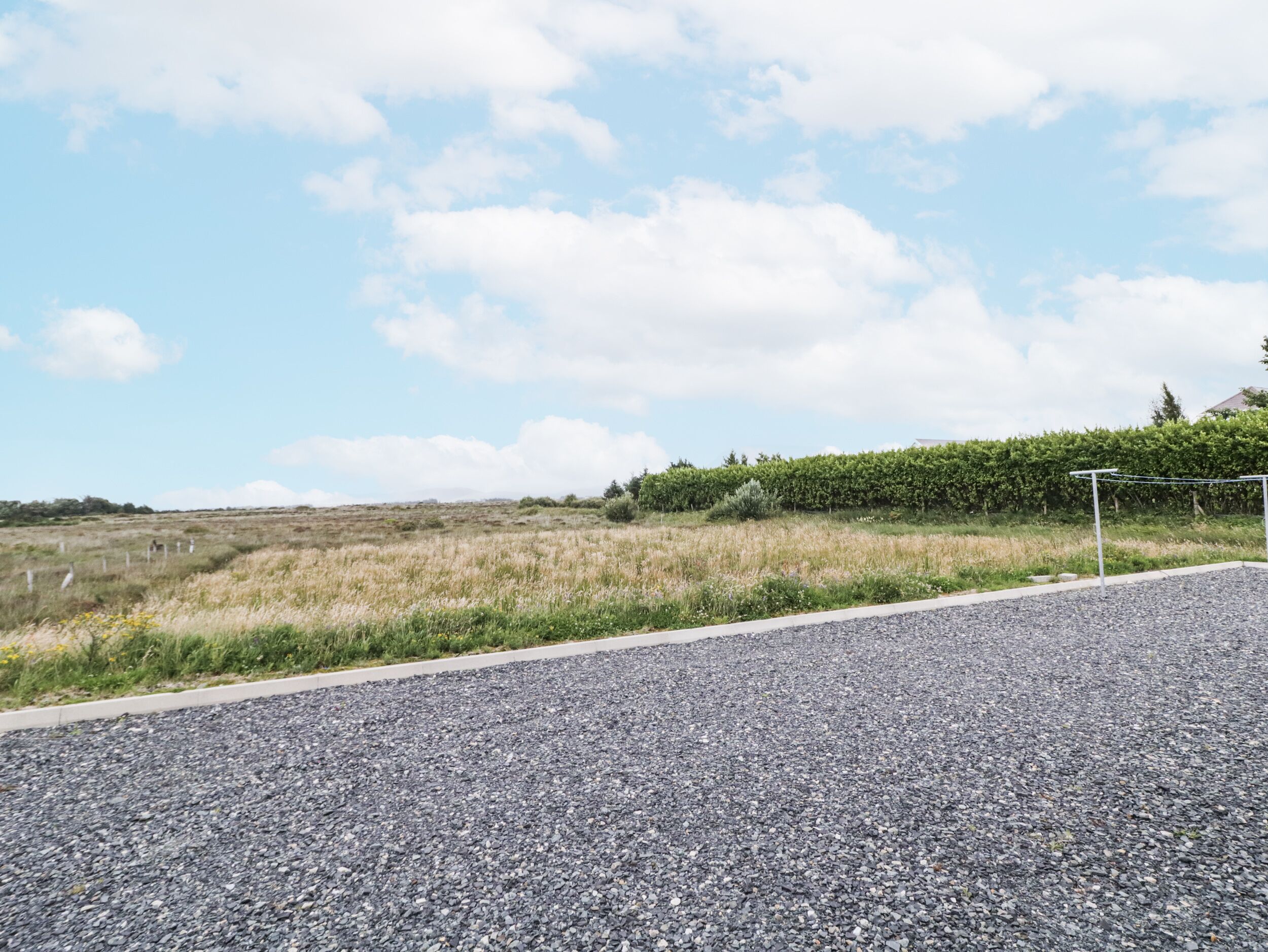 A gravel driveway leading to a grassy field with a hedge and a metal structure on the right.