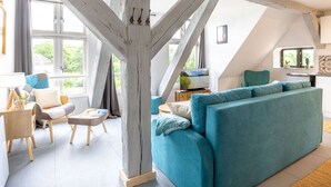 A spacious loft-style living area with exposed wooden beams, a large window offering a view of greenery, a teal sofa, and a wooden coffee table.