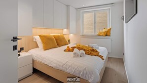 A modern bedroom with a bed, nightstands, a window with blinds, and a mounted TV.