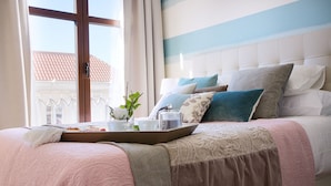 Una cama bien tendida con una bandeja de desayuno, un florero con flores y vistas al exterior a través de una ventana.