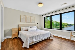 Een slaapkamer met een groot raam, een bed, een nachtkastje en een wandlamp.