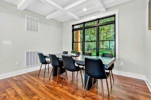 Een moderne eetruimte met een grote tafel, zes stoelen en uitzicht op een bos door grote ramen.