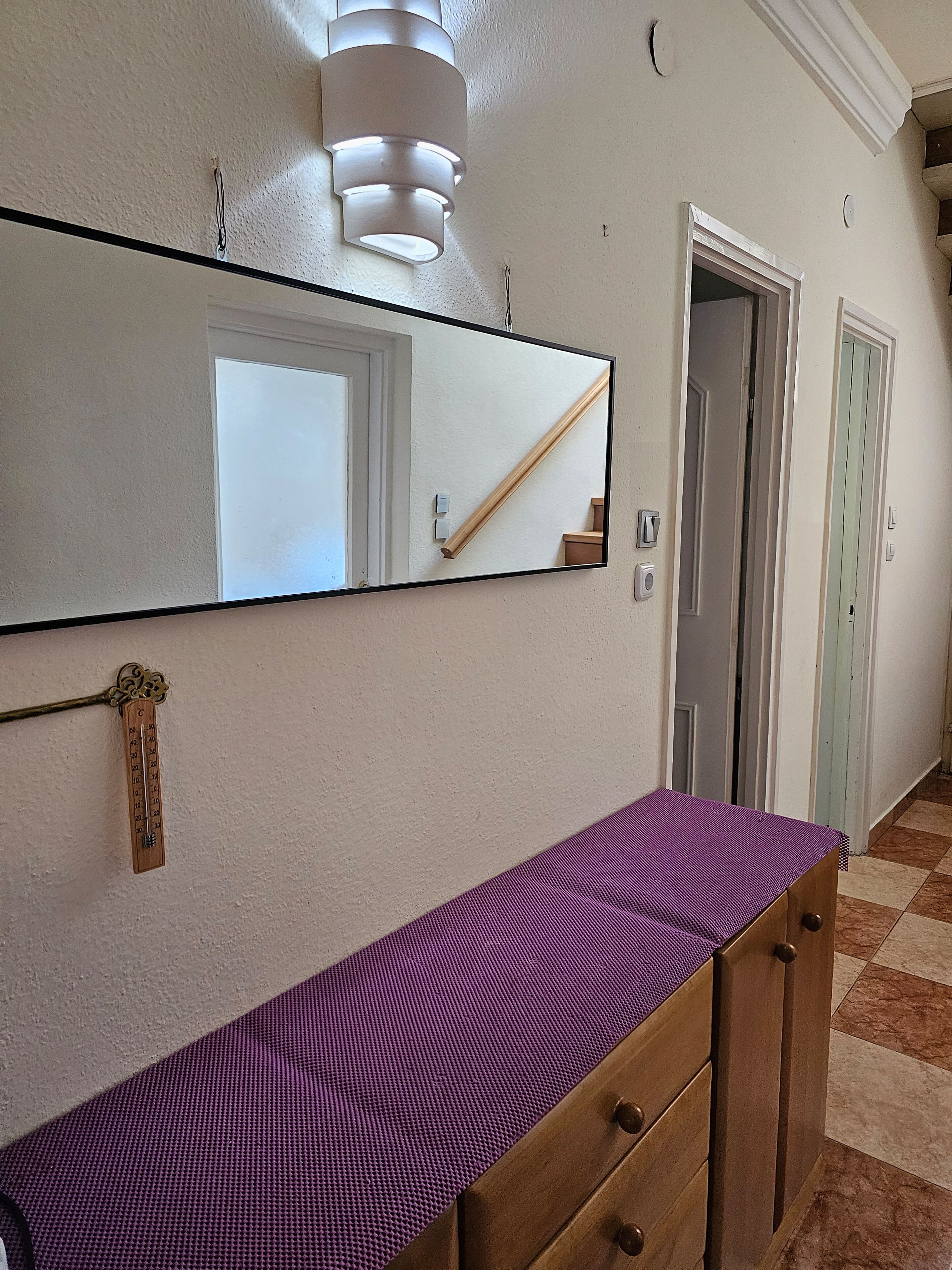 En hall med en stor spegel, en byrå i trä med lådor, en lila matta på byrån, en lampa ovanför spegeln och en dörr med en träram.