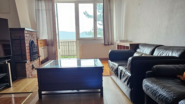 A living room with a wooden floor, a black leather sofa, a glass-top coffee table, and a fireplace. - Bright house with garden, balcony, panoramic view and fully equipped kitchen. (Pilisvörösvár)