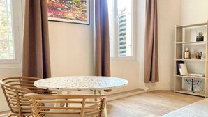 Ein Zimmer mit einem runden Tisch, Korbstühlen, einem Bücherregal und einem Fenster mit Vorhängen.