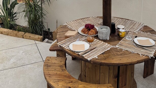 A round wooden table with a woven table runner, surrounded by benches. A thatched umbrella provides shade.
