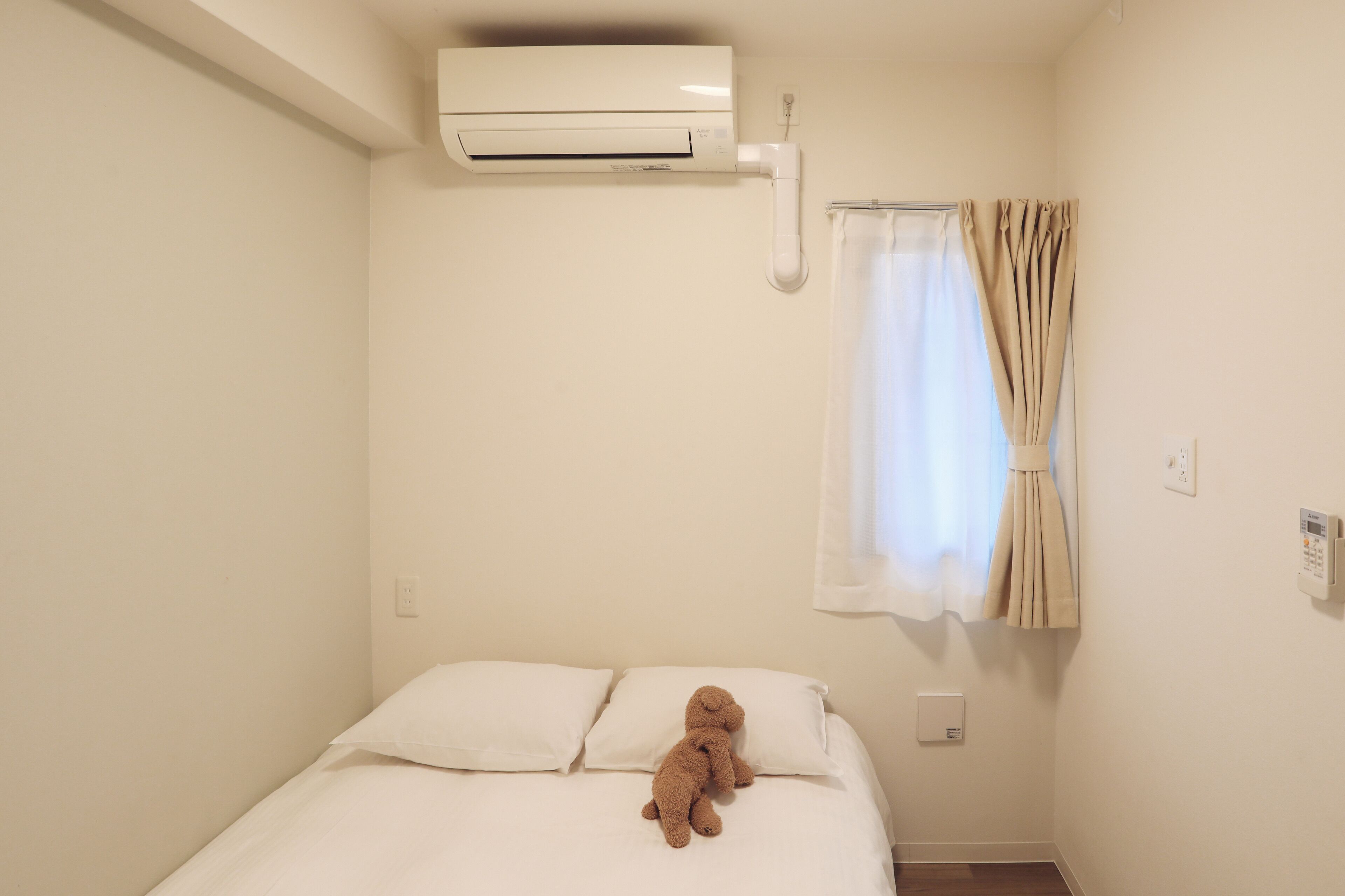 A bedroom with a bed, a teddy bear, an air conditioner, a curtain, and a thermostat.
