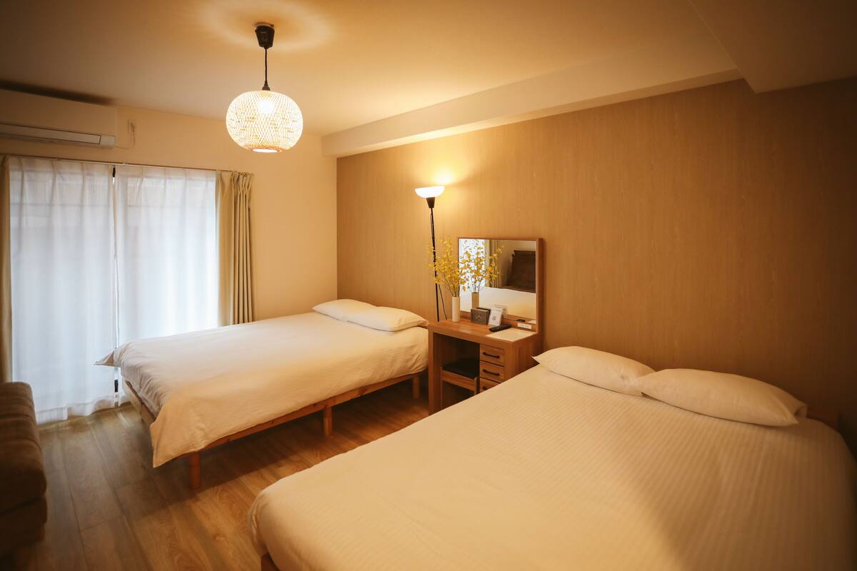 Ein Hotelzimmer mit zwei Einzelbetten, einem Schreibtisch mit Stuhl, einer Lampe und einem großen Fenster mit Vorhängen.