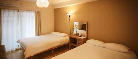 A hotel room with two single beds, a desk with a chair, a lamp, and a large window with curtains.