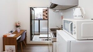 Cozinha compacta com micro-ondas, chaleira e uma pia pequena. Tem uma mesa de madeira com uma cadeira e um vaso de flores. Uma porta dá acesso a uma varanda externa.