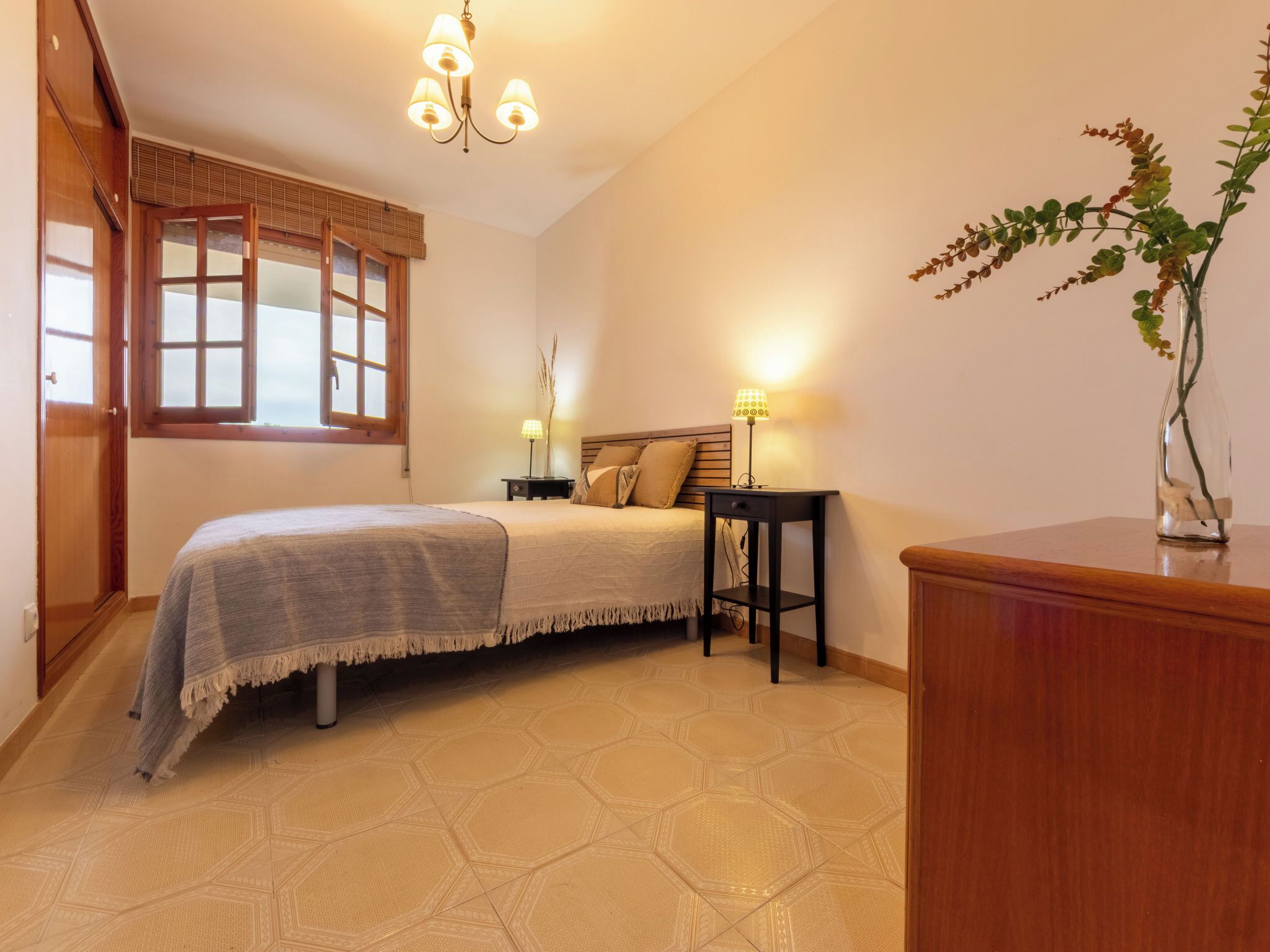 Un dormitorio con cama, mesitas de noche, lámpara, ventana con persianas y una planta.
