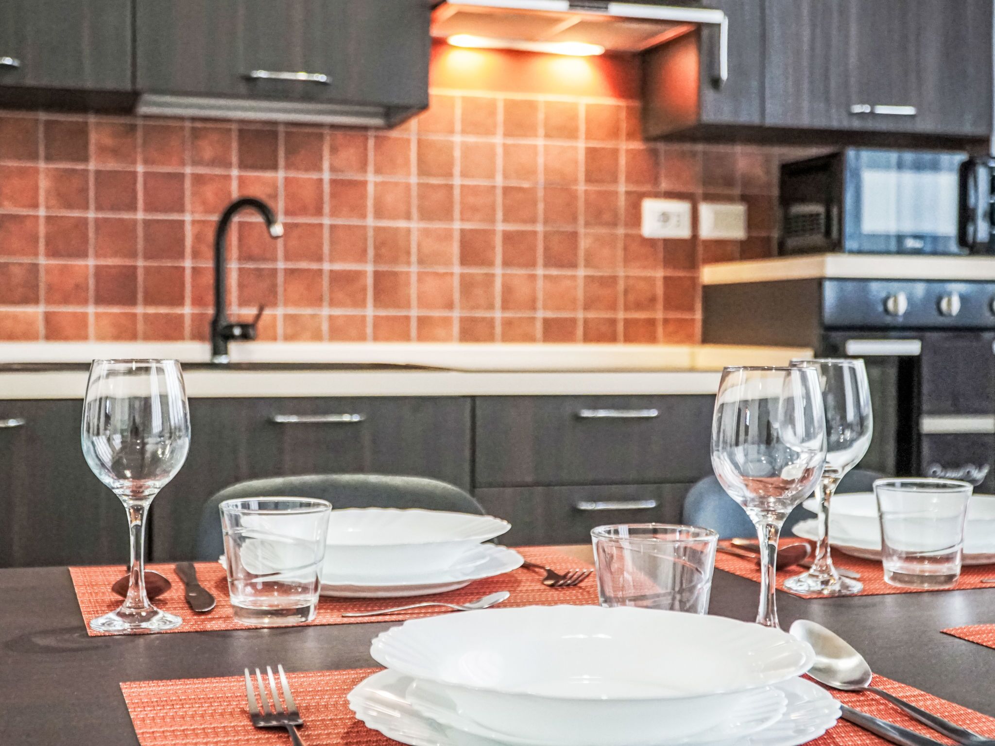 Een moderne keuken met donkere kastjes, een betegelde achterwand en een eethoek compleet met borden, glazen en bestek.