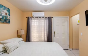 A hotel room with a bed, a ceiling fan, an air conditioning unit, a painting, a lamp, a door, and a television.