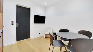 A modern room with a dark door, a flat-screen TV mounted on the wall, a round table with four chairs, and a light-colored armchair.